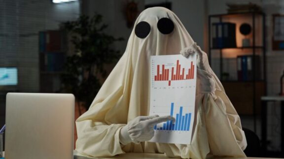 Empresario con sábana como fantasma mostrando gráficos ante un escritorio sobre el que hay un ordenador portátil