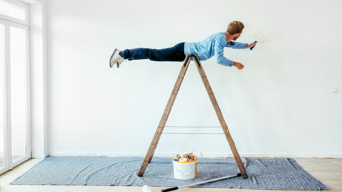 un joven con un rodillo en una mano pinta una pared haciendo equilibrio sobre una escalera
