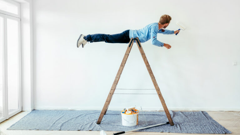 un joven con un rodillo en una mano pinta una pared haciendo equilibrio sobre una escalera