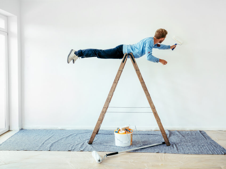 un joven con un rodillo en una mano pinta una pared haciendo equilibrio sobre una escalera