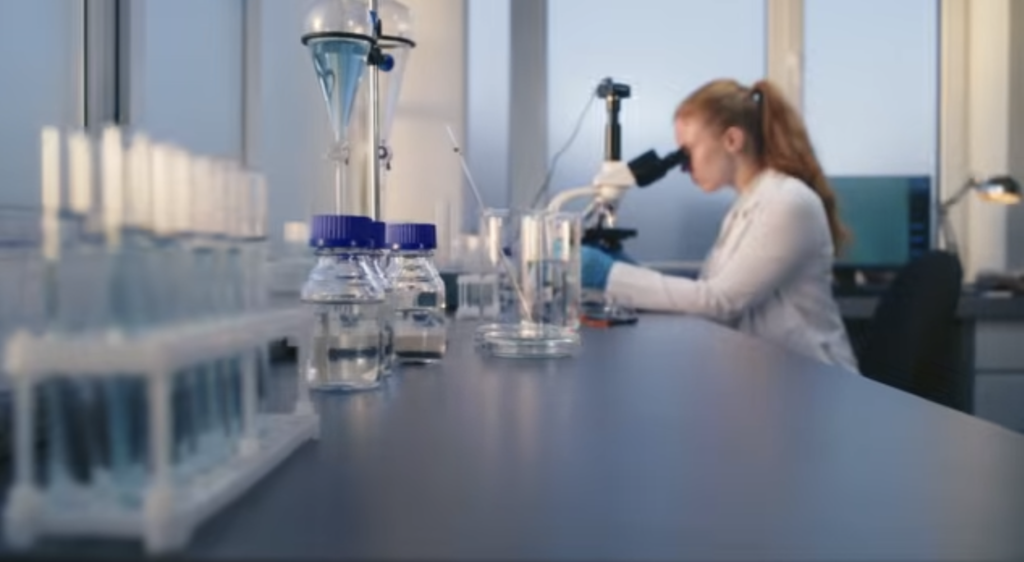 Mujer científica mirando por microscopio en un laboratorio. Imagen de Impulsa Startups (programa de impulso al emprenddimiento de la Cámara de Comercio de España)
