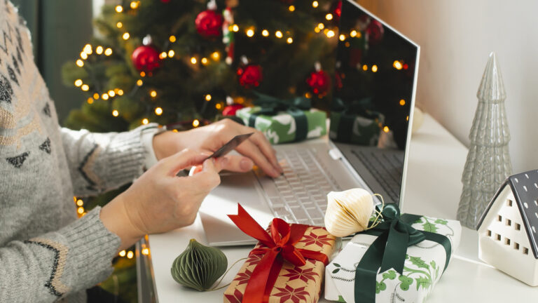 Mujer con tarjeta de pago en la mano frente a un portátil y con motivos navideños de fondo, a punto de realizar una compra en ecommerce