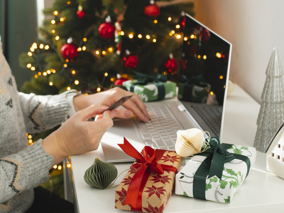 Mujer con tarjeta de pago en la mano frente a un portátil y con motivos navideños de fondo, a punto de realizar una compra en ecommerce